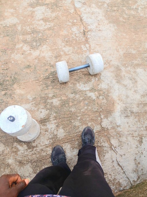In the image, a person's feet are visible while they stand next to a pair of dumbbells that have been placed upside down on the ground. The person appears to be wearing black pants and has their hands on their hips. The surrounding area seems to be a concrete or cement surface with some dirt present. There is also an object in the background, which could possibly be related to fitness equipment or exercise gear.