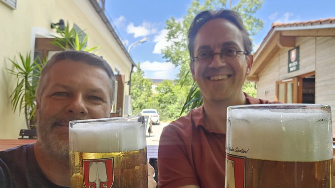 A cheerful selfie of two smiling men outdoors at what appears to be a German beer garden, with two large frothy beers in traditional glass steins in the foreground and a wooden building with green plants visible in the background under a partly cloudy blue sky.