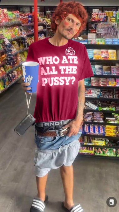 A person wearing a curly red clown-style wig, a red nose, a maroon t-shirt reading 'WHO ATE ALL THE PUSSY?', and shorts, standing in a convenience store aisle holding a large cup and a smartphone.