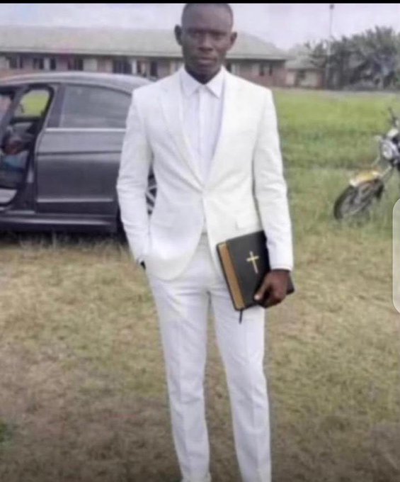 A young man wearing an all-white suit, holding a Bible with a cross on it, standing outdoors near a car and motorcycle.