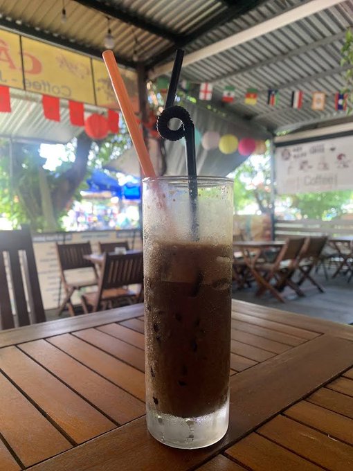 A tall glass of iced coffee with two straws sits on a wooden table at an outdoor café, with colorful lanterns and flags hanging from the ceiling in the background.
