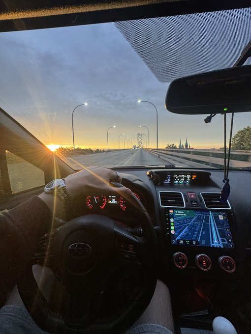 A driver's perspective from inside a Subaru during sunrise on a highway bridge, with a navigation screen and dashboard visible, heading toward Georgia as mentioned in the post. The warm morning light streaming through the windshield complements the "back on the road again" message, capturing the start of a journey.