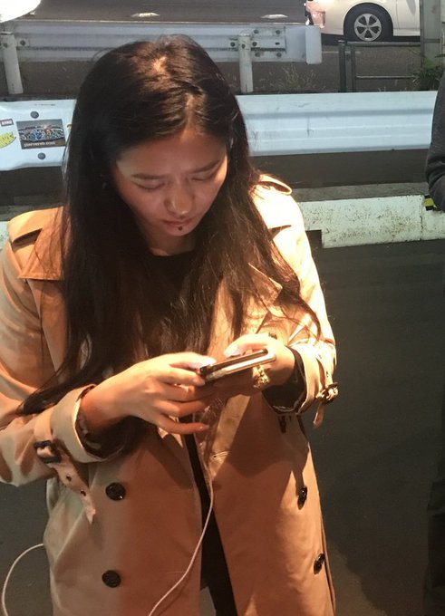 A young woman with long dark hair wearing a beige trench coat looks down at her smartphone, with a white earphone cable visible, standing near a guardrail at night.