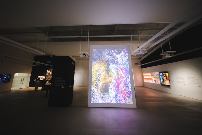 A modern art gallery displays a large illuminated freestanding screen showing swirling colorful abstract art, with smaller artworks and monitors along the surrounding walls.