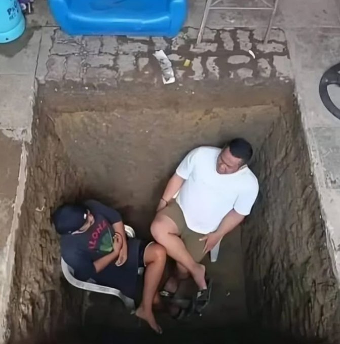 Two men sit on white plastic chairs at the bottom of a deep, narrow rectangular pit dug into the ground, with a blue inflatable pool visible at the top edge.