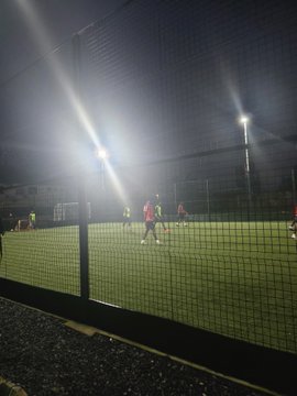 Nighttime photo of a football/soccer match being played on an artificial turf field enclosed by netting, illuminated by bright floodlights.