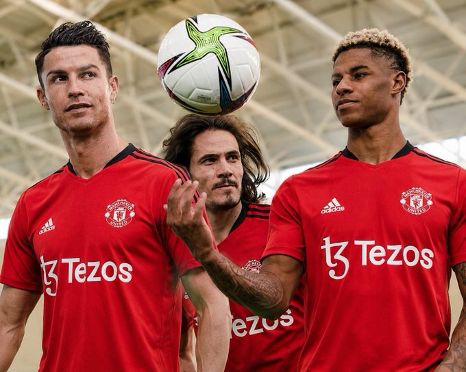 Three Manchester United football players in red Adidas kits with 'Tezos' sponsor logos pose together, with one balancing a white and green football on his hand.