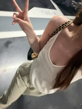 Casual selfie of a woman with long brown hair wearing a white tank top and beige cargo pants, throwing up a peace sign while standing near a white vehicle.