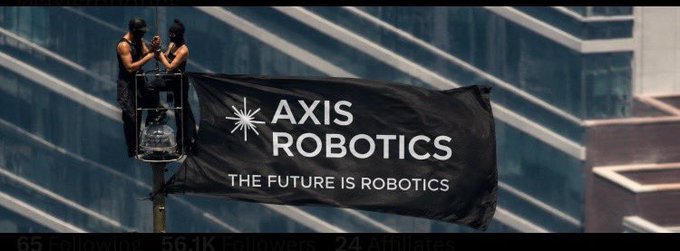 Two workers on a lift platform install a large black 'AXIS ROBOTICS - THE FUTURE IS ROBOTICS' flag on the side of a glass skyscraper.