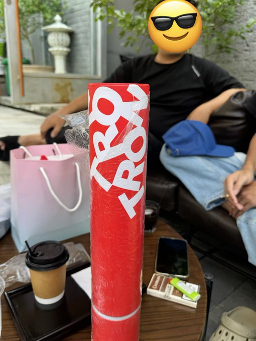 A tall red-wrapped bouquet with 'PROPOSE' text stands on a table with coffee and a phone, in front of a seated person whose face is covered by a cool sunglasses emoji.