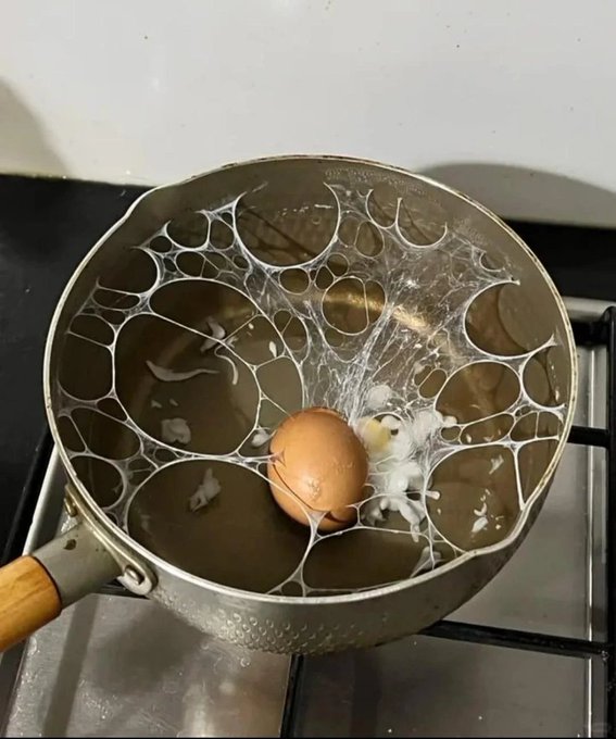 Photo of a cracked brown egg spilling yolk and white into a metal pot on a stove, with the web-like egg whites stretching across the pan's surface.