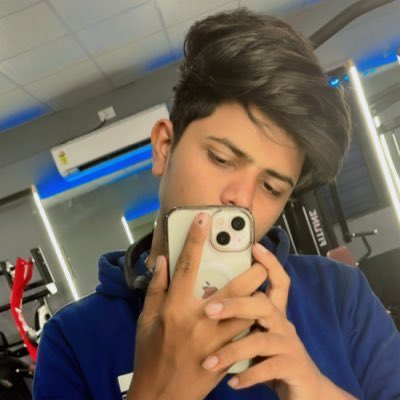 A young man with dark hair wearing a blue hoodie takes a mirror selfie with a smartphone in what appears to be a gym.