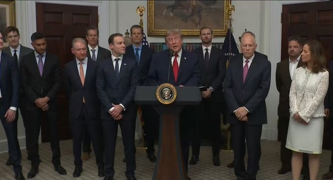 A man stands at a podium bearing the presidential seal, addressing a group of officials in suits inside an ornate room with American flags and a painting on the wall. The post suggests the man's remarks could be interpreted as bullish or bearish signals for Bitcoin, asking followers to guess his true stance.