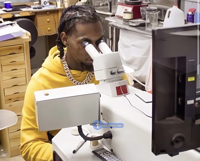 A man with braids wearing a yellow hoodie looks through a laboratory microscope in what appears to be a scientific workspace, with 'Daily Crypto vibes' watermark visible.