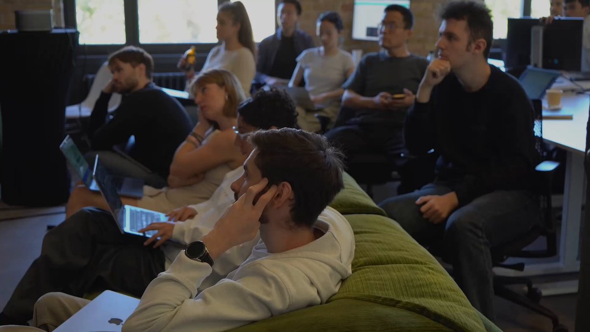 The image shows a group of people sitting on couches around each other, with some looking at their laptops while others are engaged in conversation or simply observing something happening outside the room. The scene appears to be taking place in an office setting, as there is a desk and chairs visible within the space. A laptop can also be seen placed on one of the couches. The people seem to be focused on various tasks, possibly working together or discussing ideas related to their work.