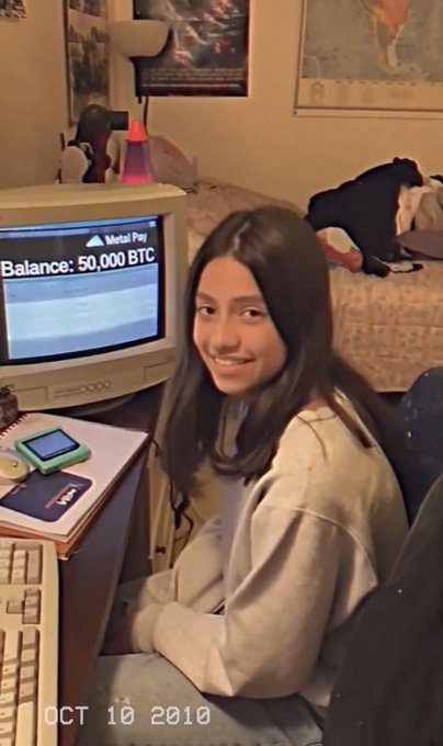 A young woman with long dark hair smiling at a vintage computer setup, with the screen displaying 'Metal Pay' and 'Balance: 50,000 BTC'. The photo is dated 'OCT 10 2010' in the bottom left corner, capturing an early Bitcoin-era moment.