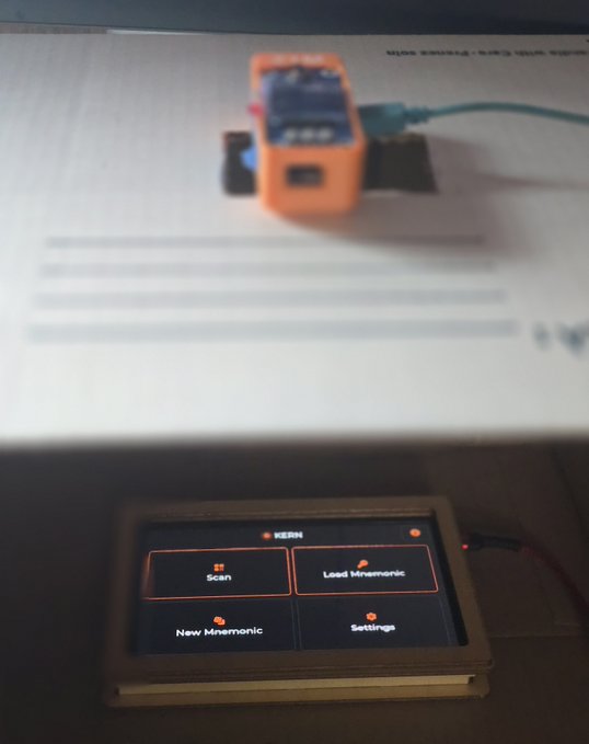 A KERN-branded device with a touchscreen displaying four options (Scan, Load Mnemonic, New Mnemonic, Settings) sits on a desk beneath a document and orange electronic device.