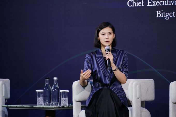 A woman with short black hair, identified as Gracy Chen, CEO of Bitget, speaks into a microphone while seated on a panel at the BRIDGE Summit, with a screen reading "Chief Executive" and "Bitget" visible behind her. She wears a striped blazer and gestures with her free hand, with water bottles on the table beside her.