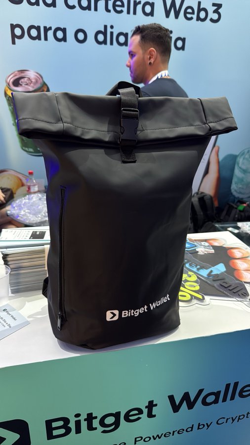 A black roll-top backpack with the "Bitget Wallet" logo sits on a display table at a promotional booth, with Portuguese text "Sua carteira Web3 para o dia a dia" visible on the background banner.