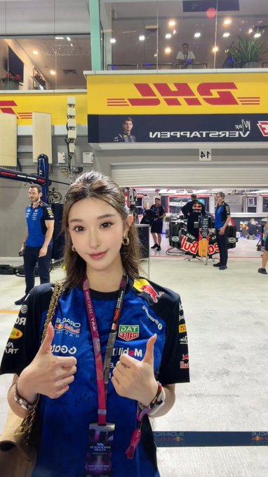 A young woman in a blue Oracle Red Bull Racing team shirt giving two thumbs up at what appears to be a Formula 1 event, standing in front of the Oracle Red Bull Racing garage area.