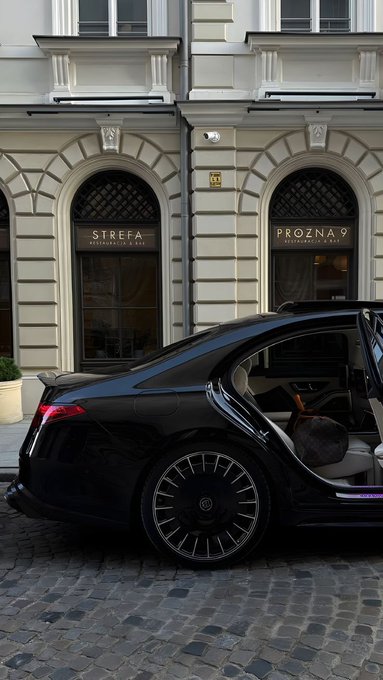 A black luxury car is parked in front of a building with arched windows labeled 'STREFA ARCHITEKCI' and 'PRÓZNA 9 APARTMENTS WAR.', parked on a cobblestone street.