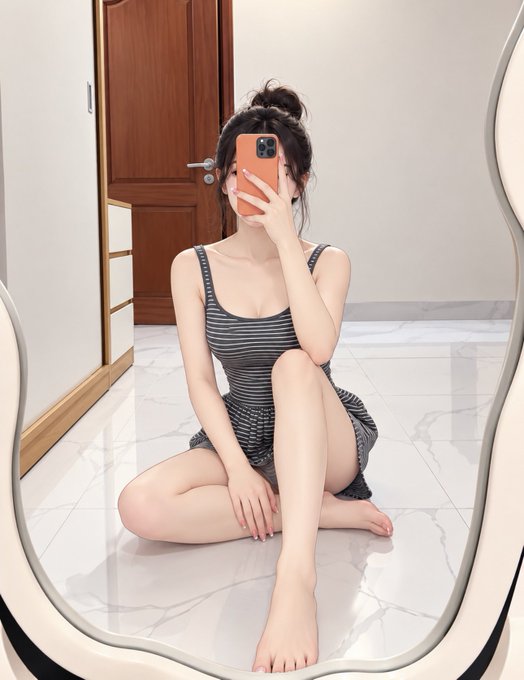 Mirror selfie of a young woman with dark hair in a bun, wearing a striped gray dress, sitting on a marble floor with an orange phone case covering her face.