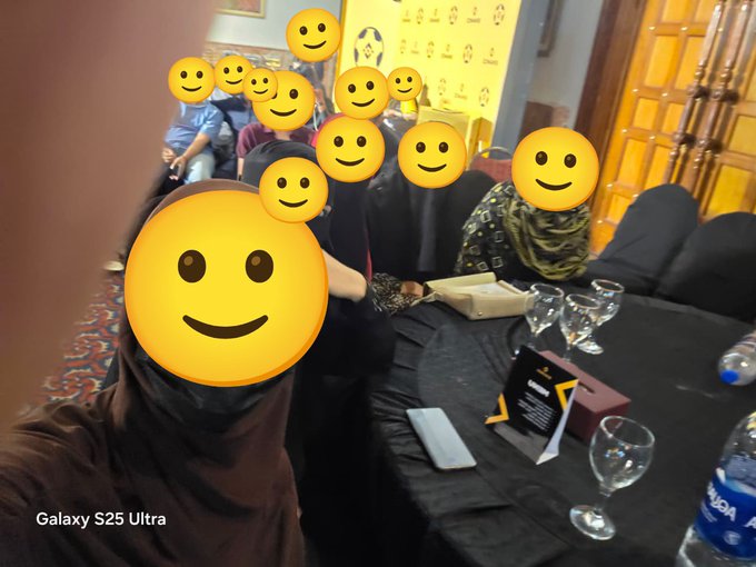 A group selfie taken with a Galaxy S25 Ultra showing people seated at a table with wine glasses and water bottles, their faces obscured by yellow smiley emojis.