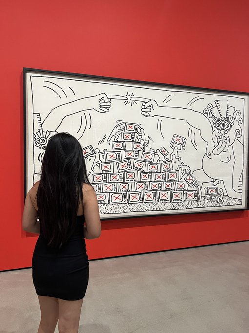 A woman with long dark hair in a black dress views a large black-and-white Keith Haring-style artwork on a red gallery wall, depicting figures with television boxes marked with X's.