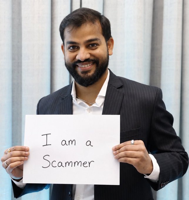 A smiling man in a dark pinstripe suit holds up a handwritten sign reading "I am a scammer," used to publicly call out an Indian crypto platform that allegedly stole ₹27,500 from him and refused to resolve the issue.