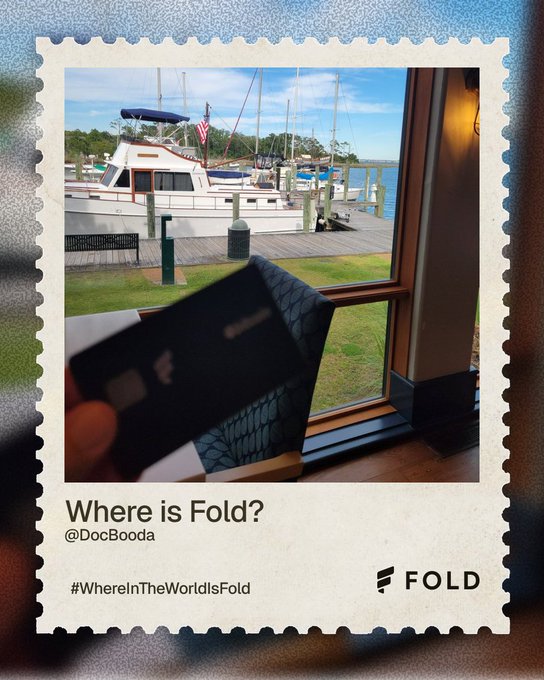 A Fold-branded card is held up to a window overlooking a marina with boats and an American flag, with the caption "Where is Fold? @DocBooda" and hashtag "#WhereInTheWorldIsFold" displayed below—fitting the post's prompt asking followers to guess the boat's destination before a humorous "accidental boating accident."