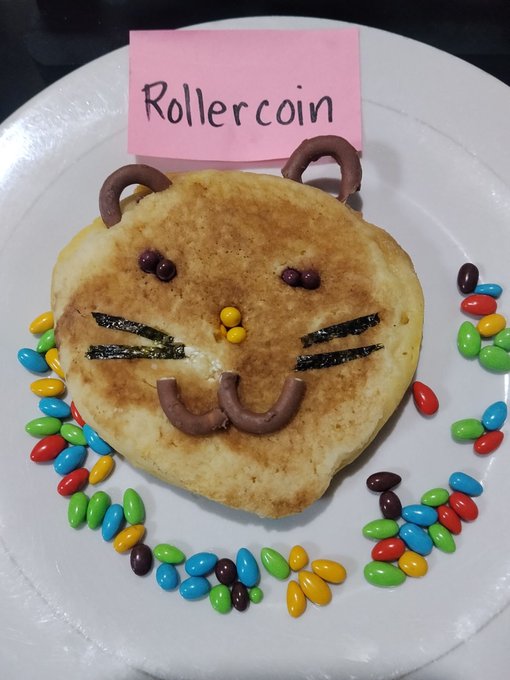 A creative pancake art on a white plate depicting a cartoon bear face with chocolate features, surrounded by colorful jelly beans, with a pink sticky note labeled 'Rollercoin'.