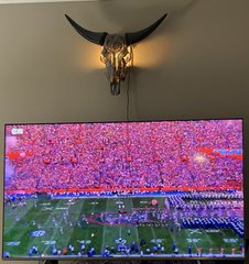 A bull skull sconce light glows warmly above a TV displaying a packed college football stadium during a game broadcast, reinforcing the "It's on!" excitement with a classic game-day setup.
