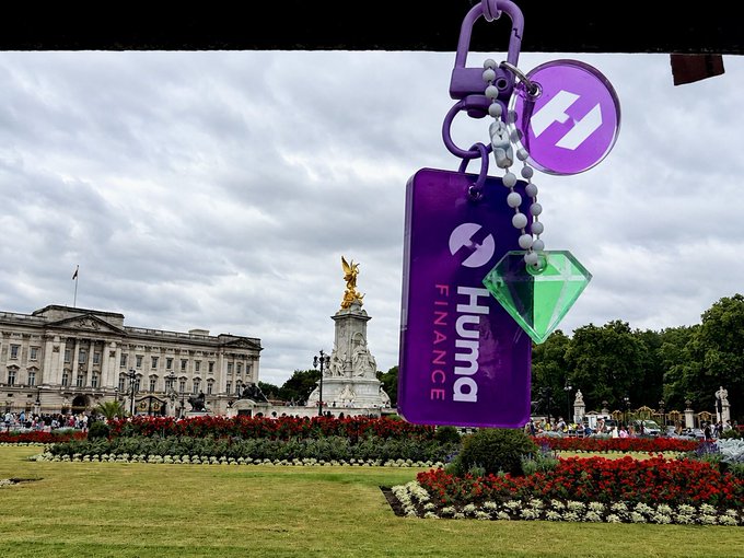 A Hum Finance promotional keychain with the purple logo and a green gem charm hanging in front of Buckingham Palace and the Victoria Memorial in London.