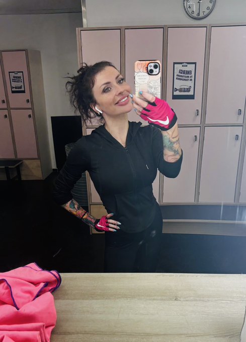 A woman in a black athletic jacket and pink Nike gloves takes a mirror selfie with a phone in a locker room, with towels and a clock visible in the background.