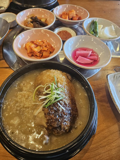A Korean meal featuring a bowl of braised short rib soup with green onion garnish, surrounded by various banchan side dishes including kimchi and pickled radish.