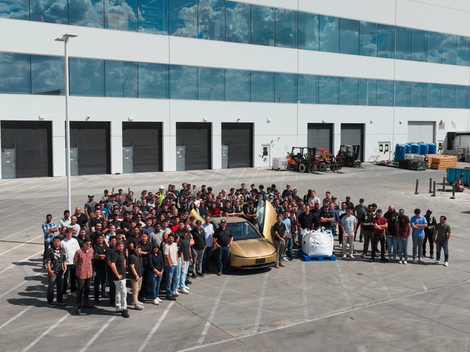 A large group photo of a company team gathered outside a glass-windowed industrial building, posing around a gold-colored sports car and construction vehicles.