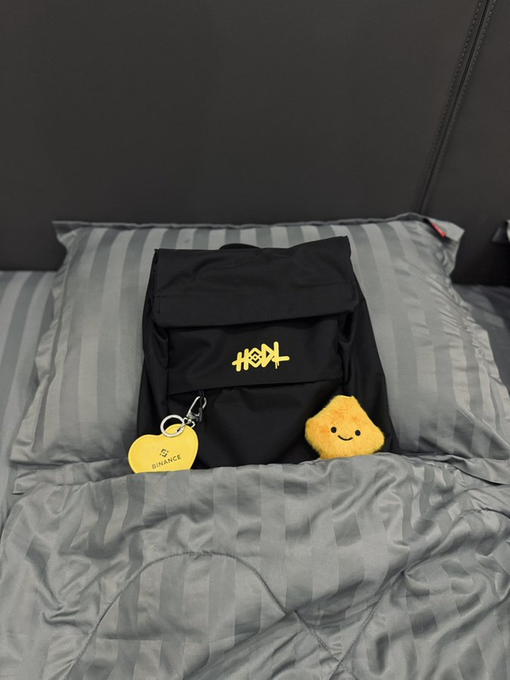 A black backpack with a gold graffiti-style logo resting on a gray striped bed, accompanied by a yellow round keychain with 'BINANCE' text and a smiling star-shaped plush toy.