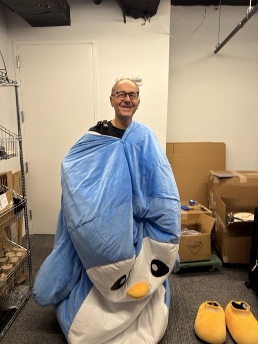A smiling man wearing glasses and wrapped in a blue penguin sleeping bag in what appears to be a storage room with boxes and metal shelving.