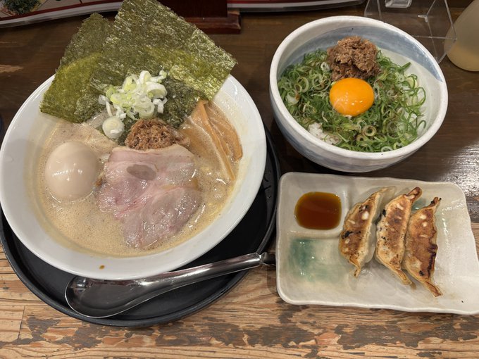 A spread of Japanese cuisine featuring a bowl of ramen with chashu pork, nori, and a soft-boiled egg, accompanied by a rice bowl topped with raw egg yolk and green onions, plus a plate of gyoza with dipping sauce.