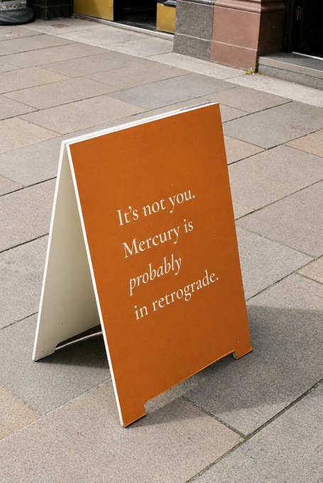 An orange A-frame sandwich board sitting on a sidewalk reads "It's not you. Mercury is *probably* in retrograde." The whimsical sign humorously reflects the post's embrace of a single astrological indicator—Mercury retrograde—as the go-to excuse for trading misfortunes, perfectly capturing the mood of relying on cosmic explanations instead of traditional market analysis.