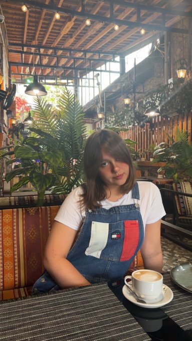 A young woman with brown hair wearing denim overalls and a white t-shirt sits at a table with a latte in a cafe decorated with lush green plants and string lights.