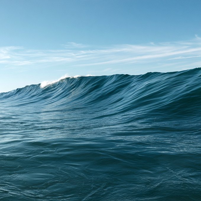 A minimalist photograph of a deep blue ocean wave curling under a clear light blue sky with wispy clouds.