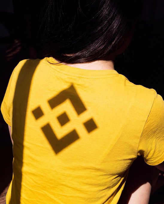 A person wearing a yellow t-shirt is photographed from behind, with sunlight casting a Binance logo-shaped shadow across their back. The image cleverly ties into the post's caption by showing how natural sunlight creates the iconic Binance symbol, turning a simple sunny weekend into a branded moment.