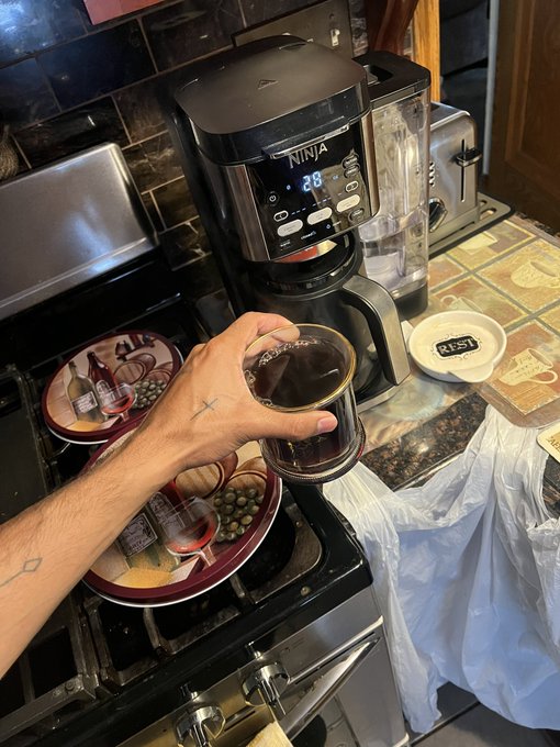 A hand reaching toward a Ninja coffee maker on a kitchen counter, holding a glass of dark liquid, with various food packages and bowls visible on the stove.