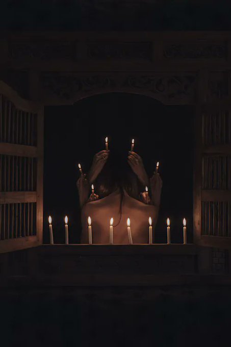 A dark, atmospheric scene showing a figure from behind surrounded by lit candles arranged in a row on a ledge, with additional candles appearing to be held by the figure, viewed through an ornate wooden window frame.