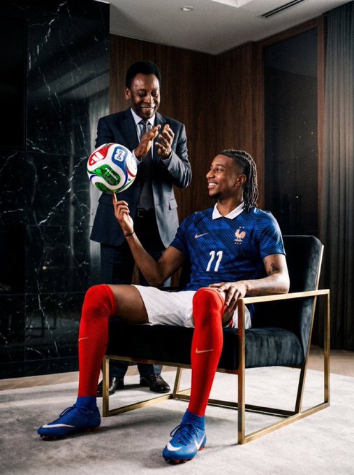 Two men, one standing in a suit and another seated wearing a France national team jersey (#11) spinning a soccer ball on his finger, pose for a photo in a modern room.