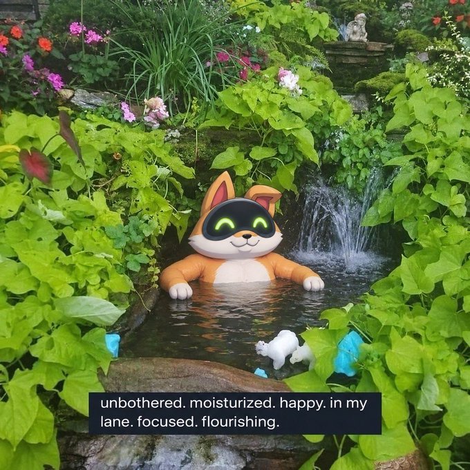 An orange and white cat-like robot toy sits in a small garden pond beneath a tiny waterfall, accompanied by a sign reading 'unbothered. moisturized. happy. in my lane. focused. flourishing.'