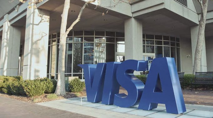Large blue "VISA" letters stand outside the entrance to one of Visa's corporate buildings. The image illustrates the article about Visa launching an internal stablecoin platform in partnership with the Open Standard stablecoin OUSD.