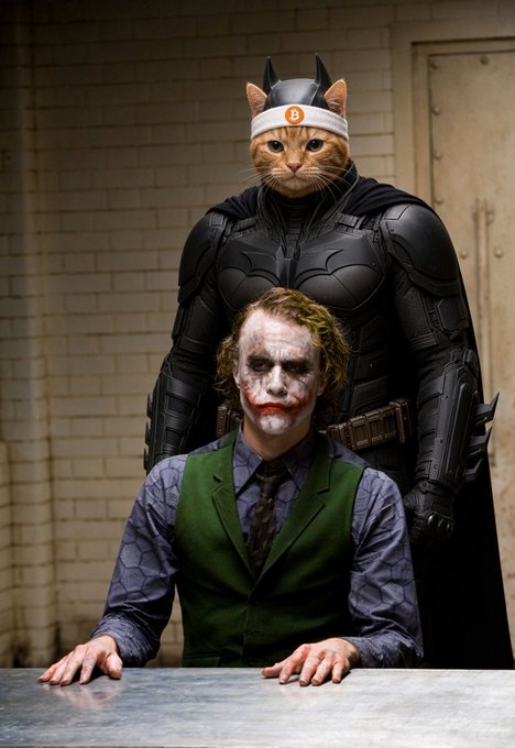 A surreal scene featuring a person with Joker-like makeup sitting at a table, while a figure in a Batman suit with a cat's head (wearing a headband with a 'B' logo) stands behind them.