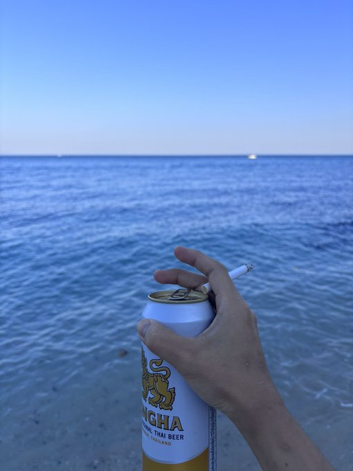 A hand holds a can of 'Singha' Thai beer and a lit cigarette against a scenic backdrop of the ocean and clear blue sky.